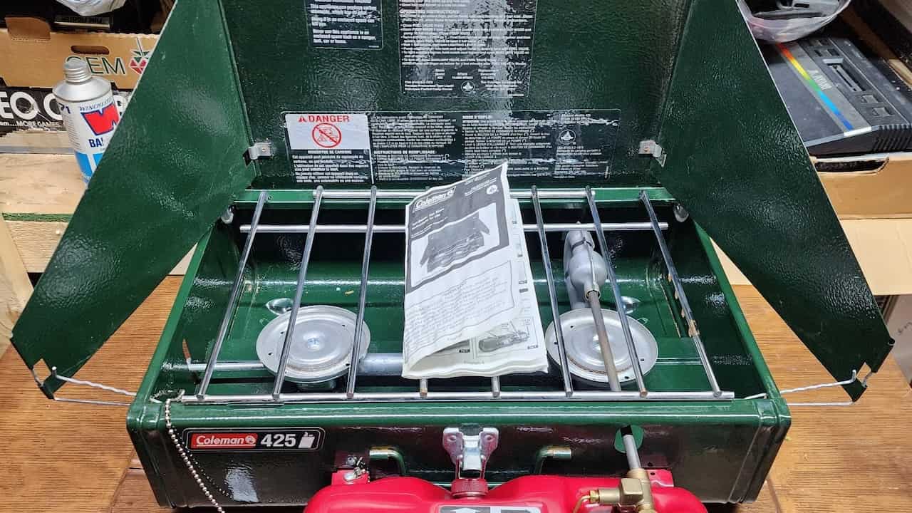 Coleman 425 two-burner camping stove with a red fuel tank and instruction manual, placed on a wooden surface in a garage or workshop