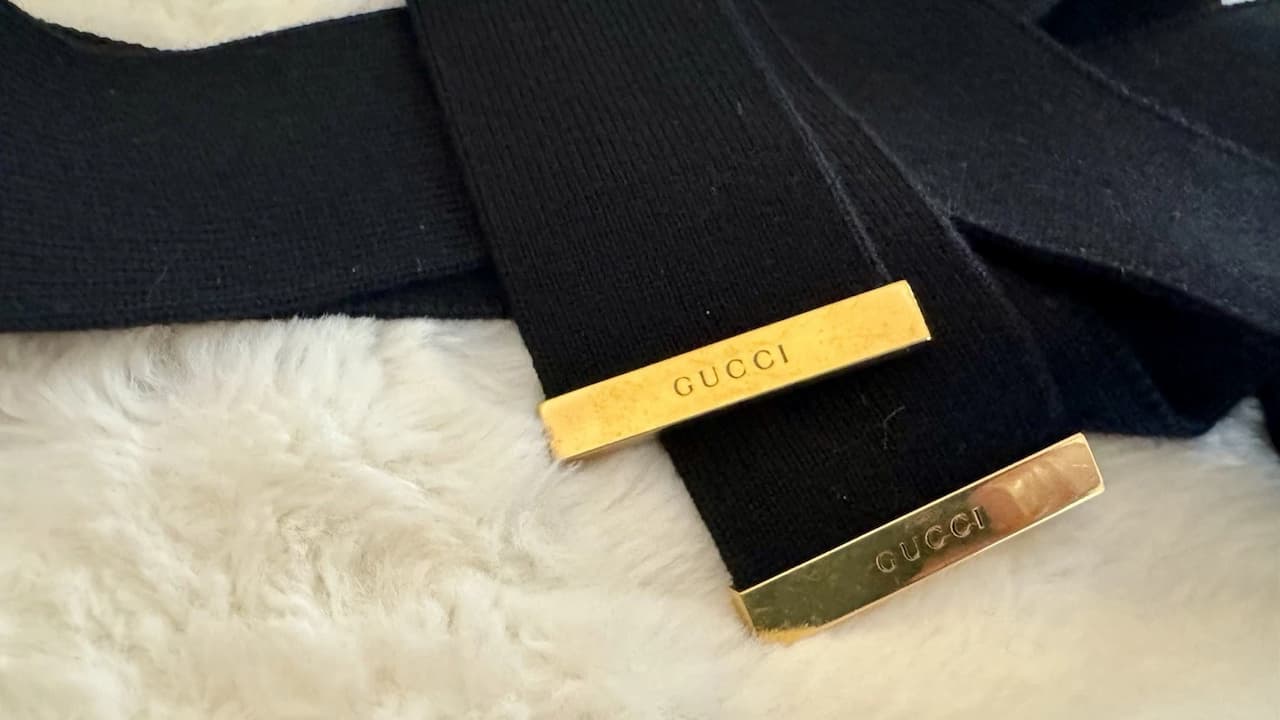 Close-up of a black fabric accessory with two gold rectangular tags engraved with the word "GUCCI," resting on a soft white fur surface