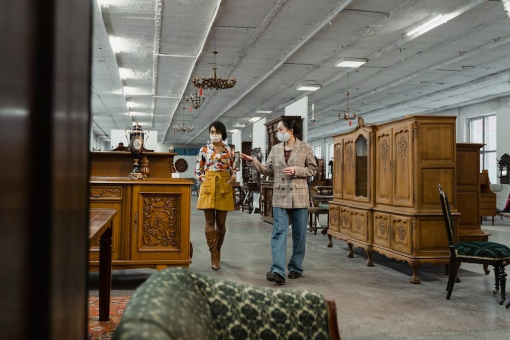 Two women wearing face masks walk through a large indoor estate sale, browsing antique wooden furniture and vintage decor under a ceiling lined with chandeliers