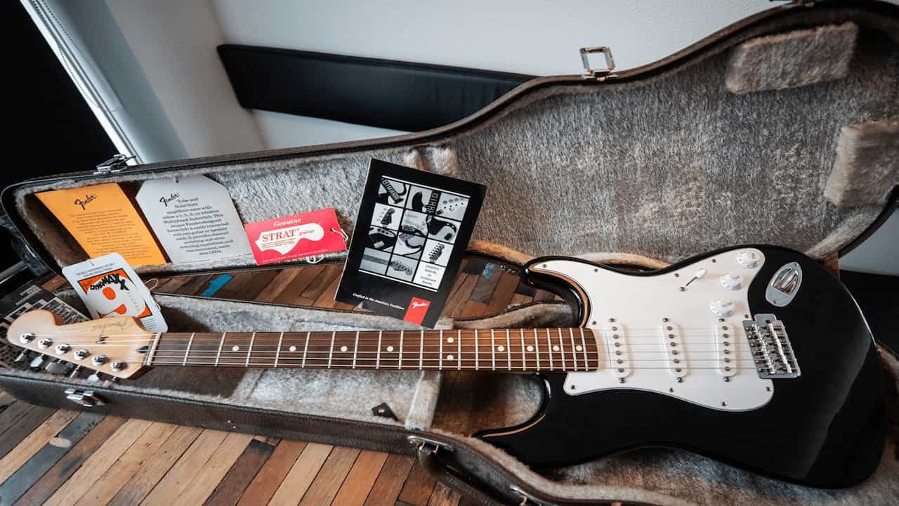 Black Fender Stratocaster electric guitar with white pickguard placed inside a grey plush-lined hard case, accompanied by tags and manualss