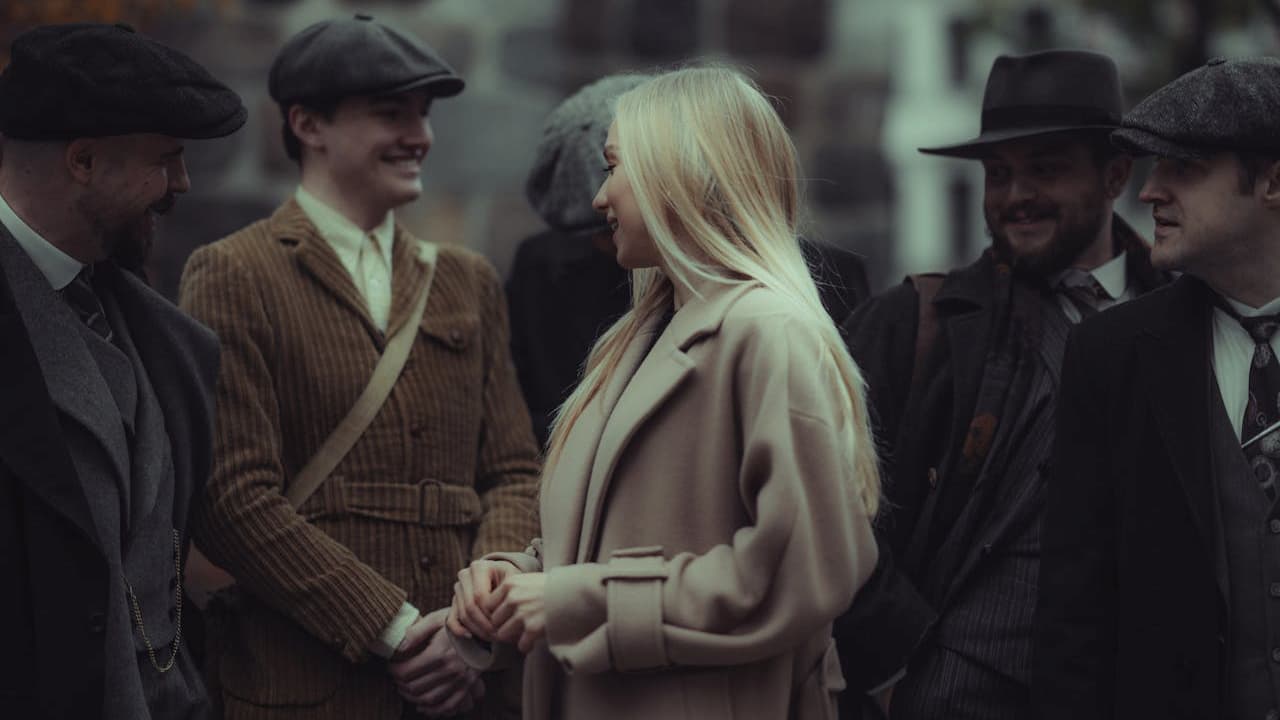 Group of people dressed in vintage-inspired clothing, including newsboy caps, trench coats, and tailored suits, standing and smiling in conversation on a street with a stone building in the background
