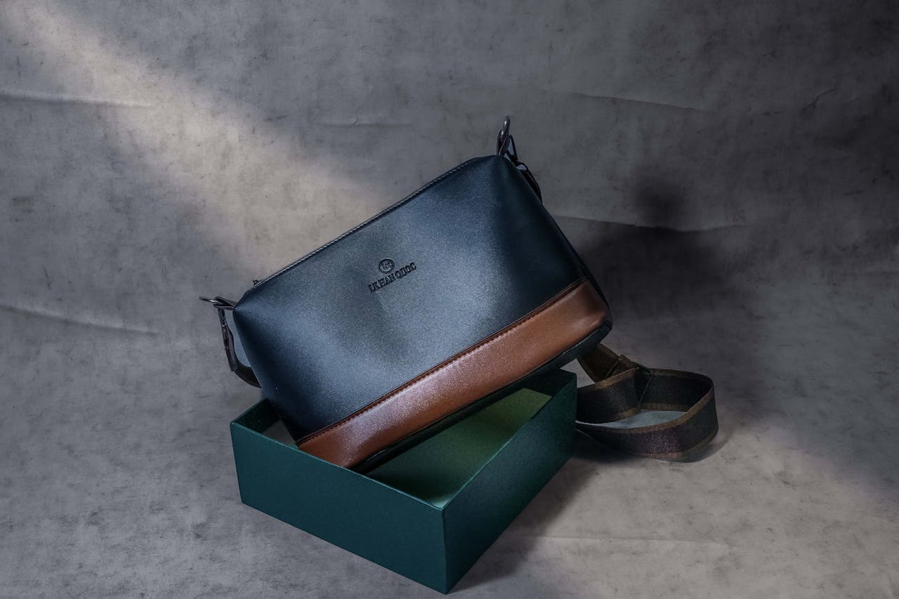 Black and brown leather crossbody bag with an adjustable strap, displayed partially inside a green box on a grey textured surface