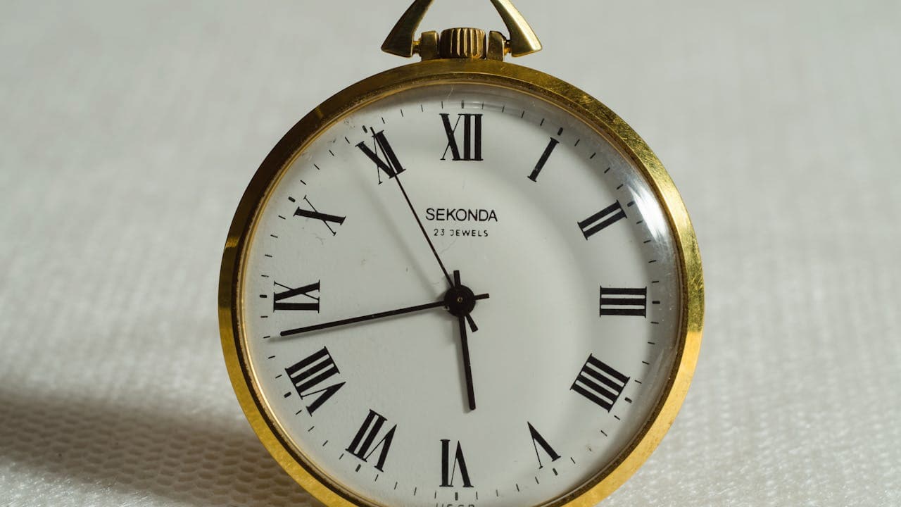 Gold-framed pocket watch, white face, black Roman numerals distorted and melting downward, black hour and minute hands, set against a light gray textured background