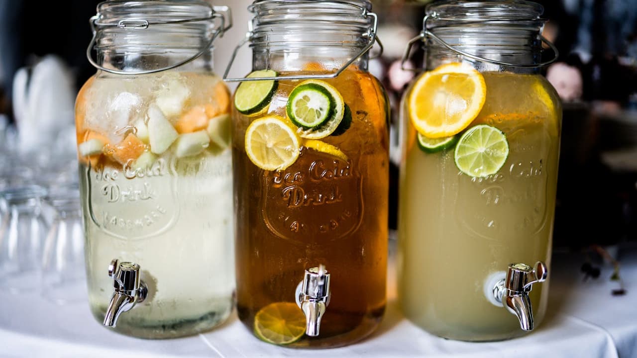 Three glass beverage dispensers with metal spouts, filled with fruit-infused drinks, displayed on a white tablecloth