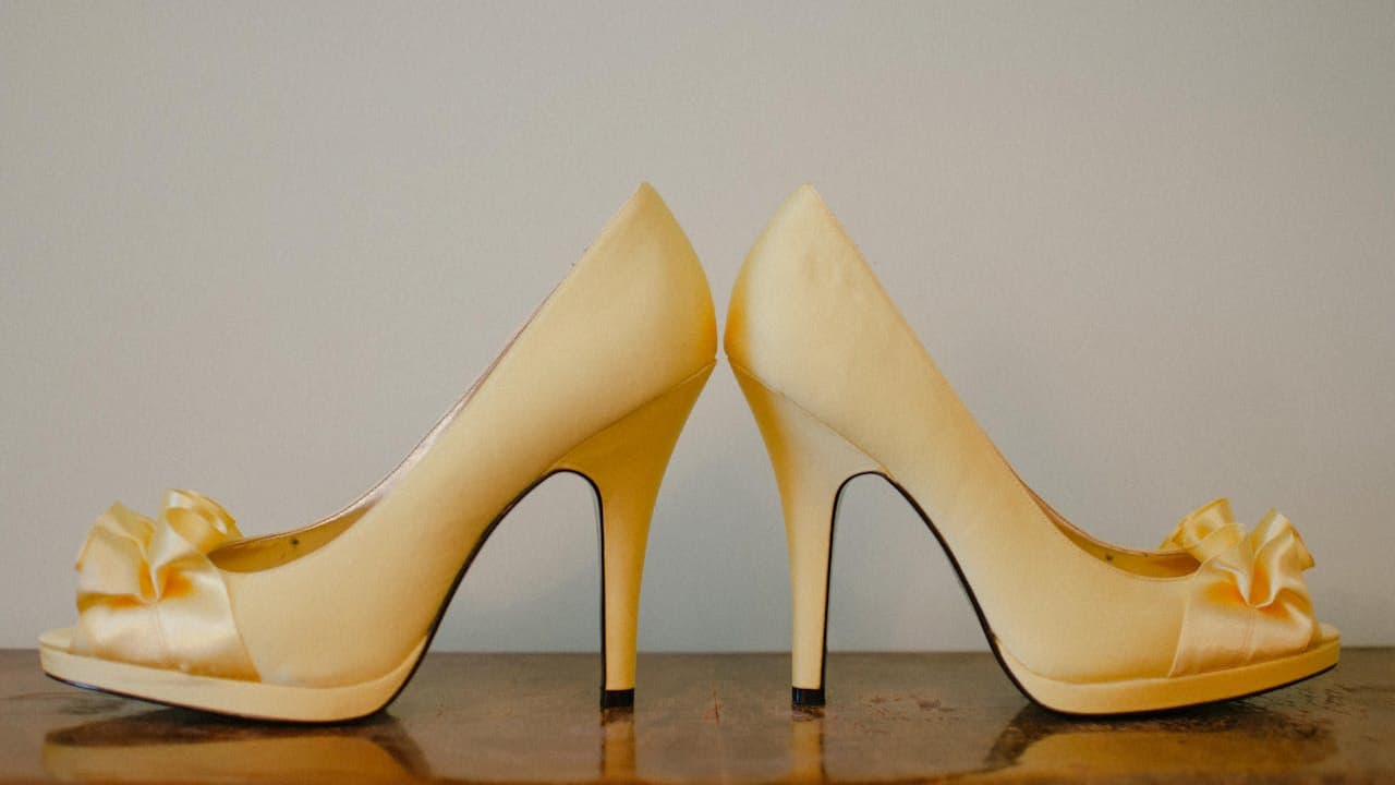 Beige high-heeled shoes, glossy finish, decorative bows on toes, displayed heel to heel, placed on a wooden surface, neutral background