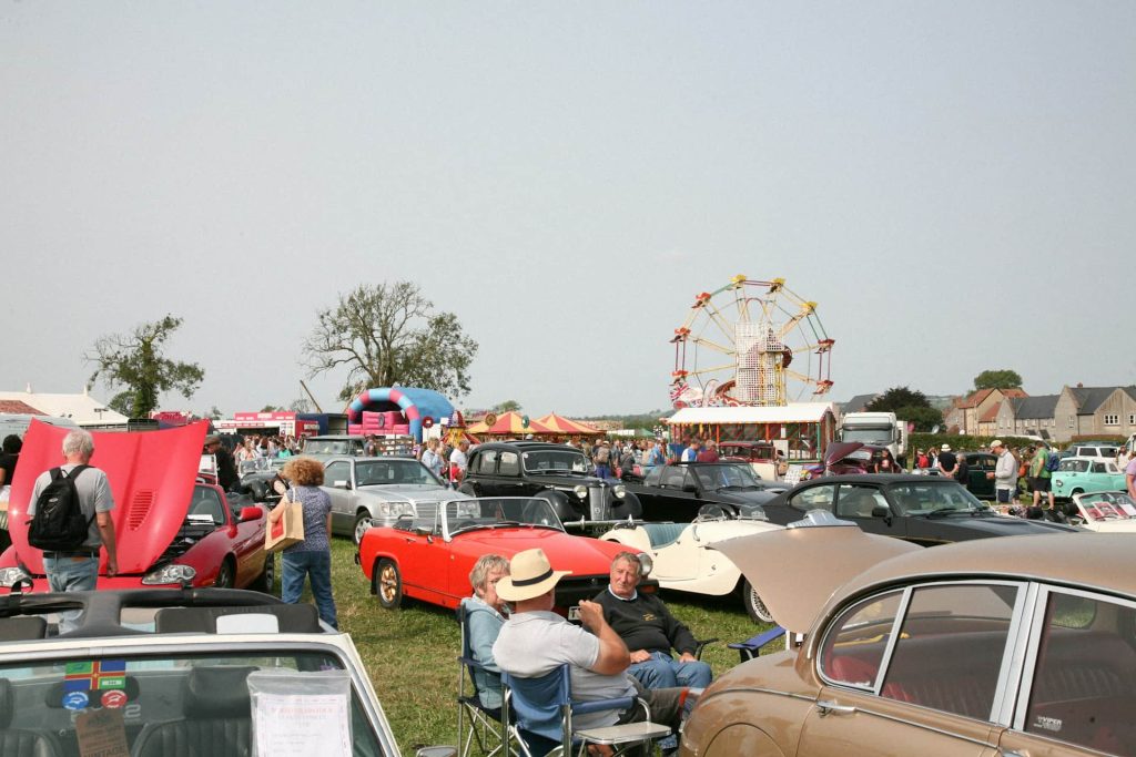 A lively country fair with vintage cars lined up on display. People are strolling, admiring the cars, and enjoying the festivities, which include a Ferris wheel, food stalls, and a bouncy castle in the distance. The atmosphere is vibrant and full of summer fun.