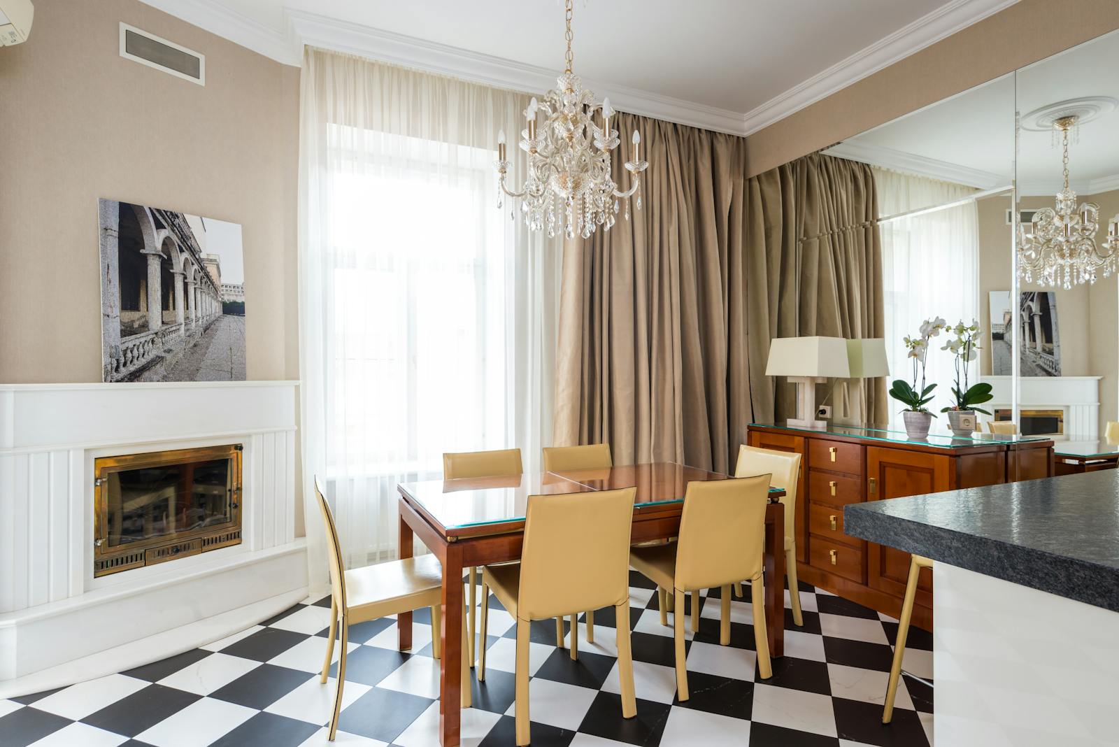 Dining table with chairs placed near window covered with curtains in kitchen with classic chandelier in apartment