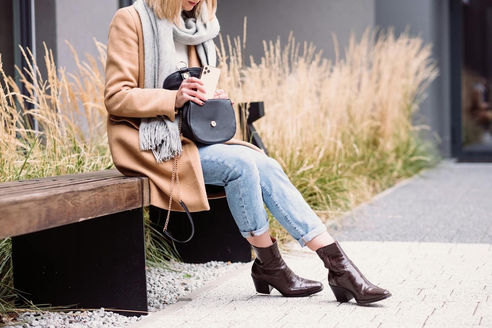 Elegant woman in a coat and scarf using her phone on a bench outdoors in Warsaw.