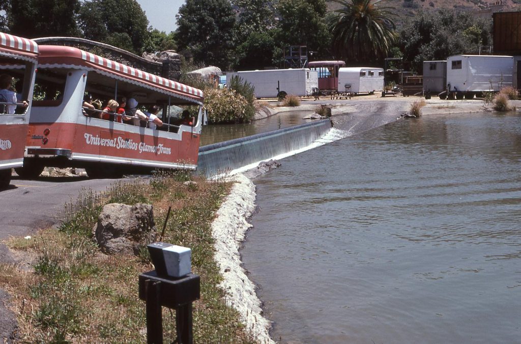 A Universal Studios tour tram crosses a scenic waterway with tourists enjoying the view.