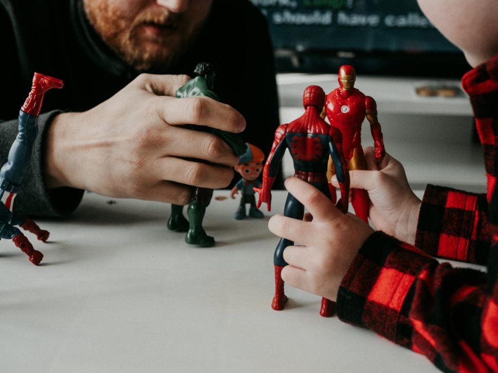 A father and son bond while playing with superhero action figures indoors.