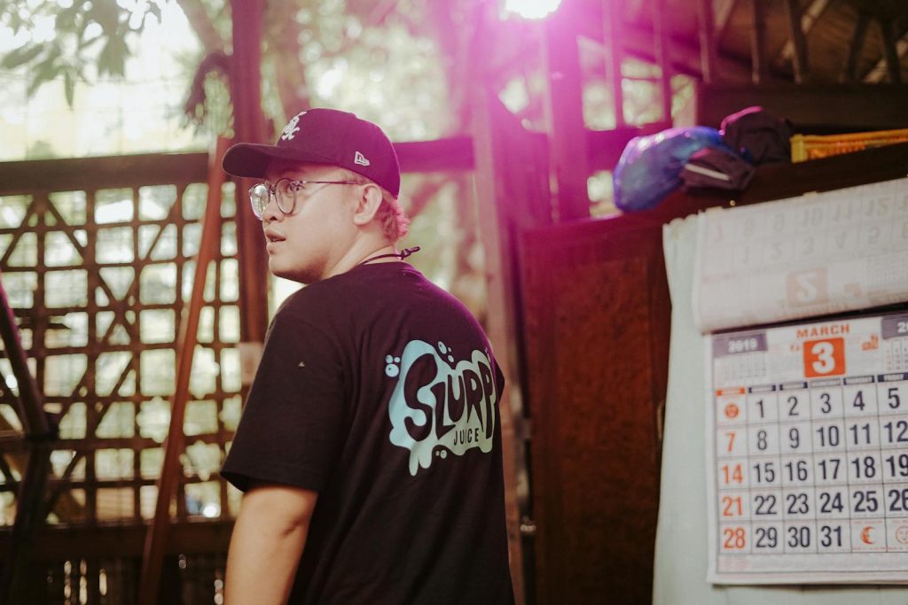 A casual young man wearing a cap and glasses stands indoors beside a wall calendar.