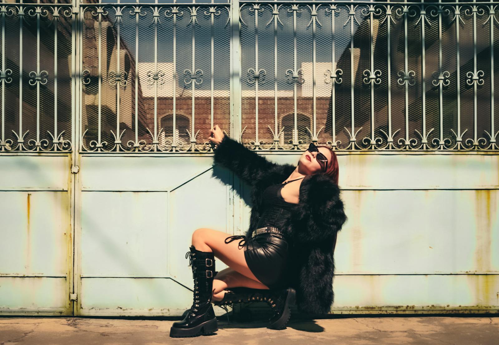 Stylish woman in leather and fur poses in front of an ornate gate in urban sunshine.