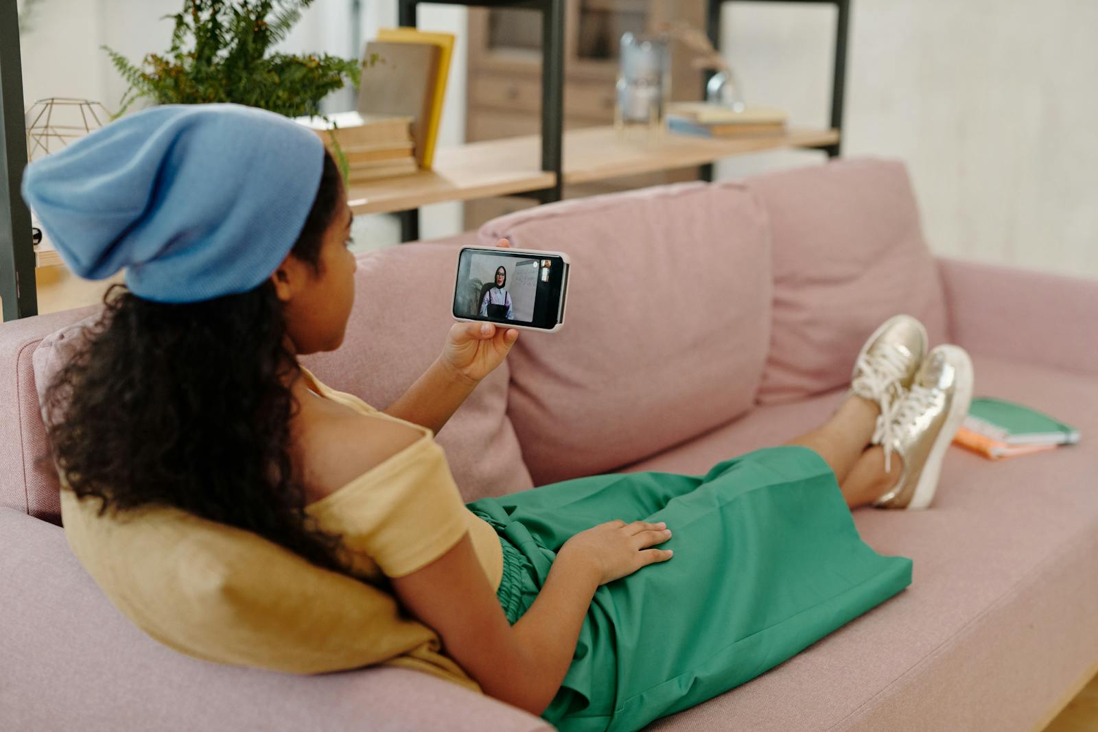 Young woman in casual attire enjoying a video call on her smartphone while lounging on a pink sofa indoors.