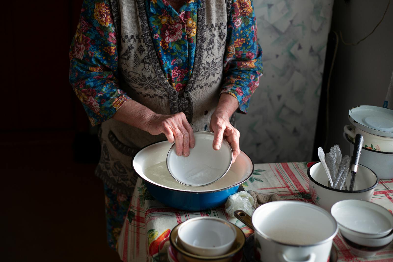 An elderly woman washes dishes by hand, capturing a timeless kitchen scene.
