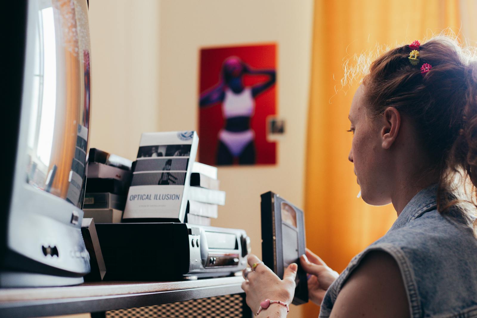 A young woman in a room holding a VHS tape, surrounded by retro equipment and decor.