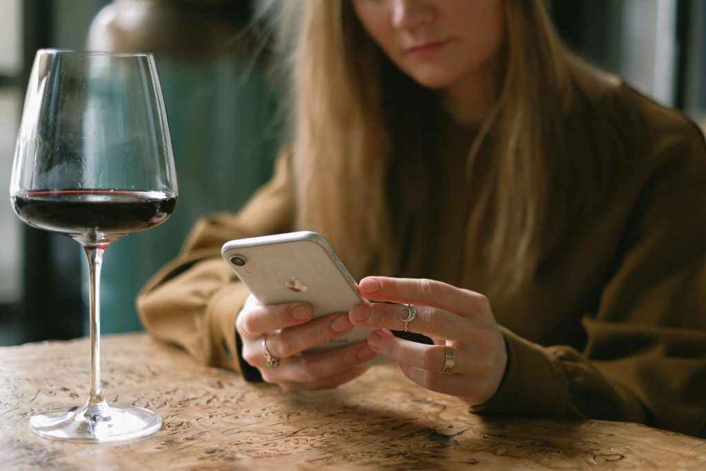 A woman is relaxing indoors, using her smartphone with a glass of red wine on the table.