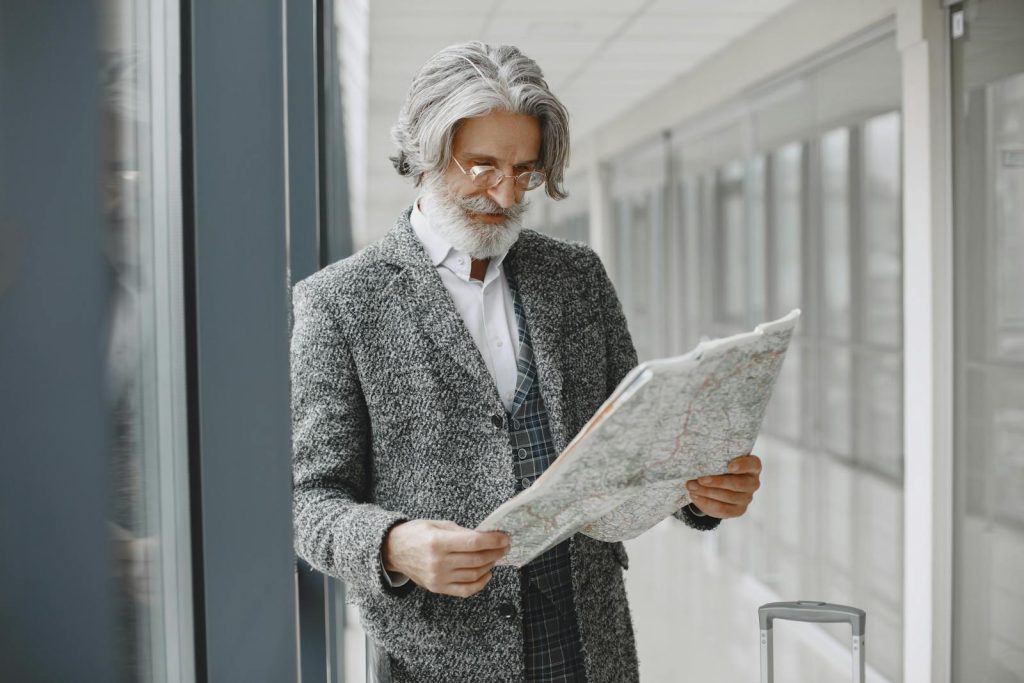 Elderly man with suitcase reads map in a bright airport corridor, planning his journey.