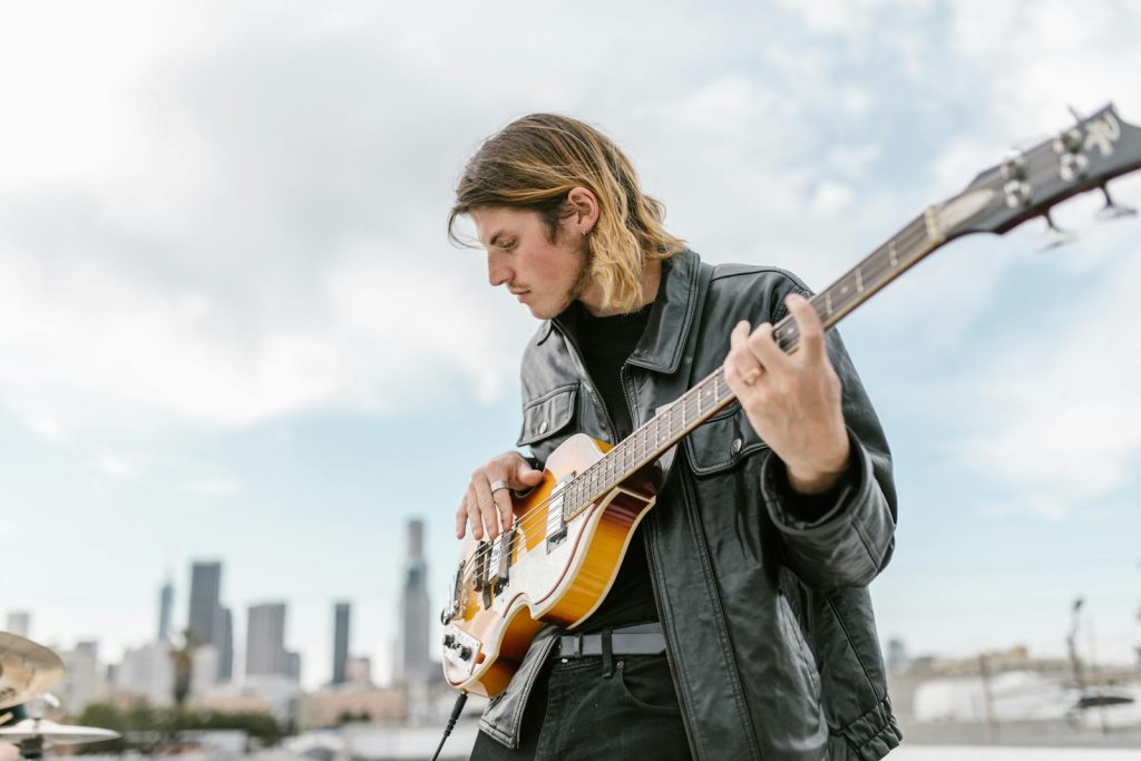 A young musician plays an electric bass guitar with a city skyline in the background.