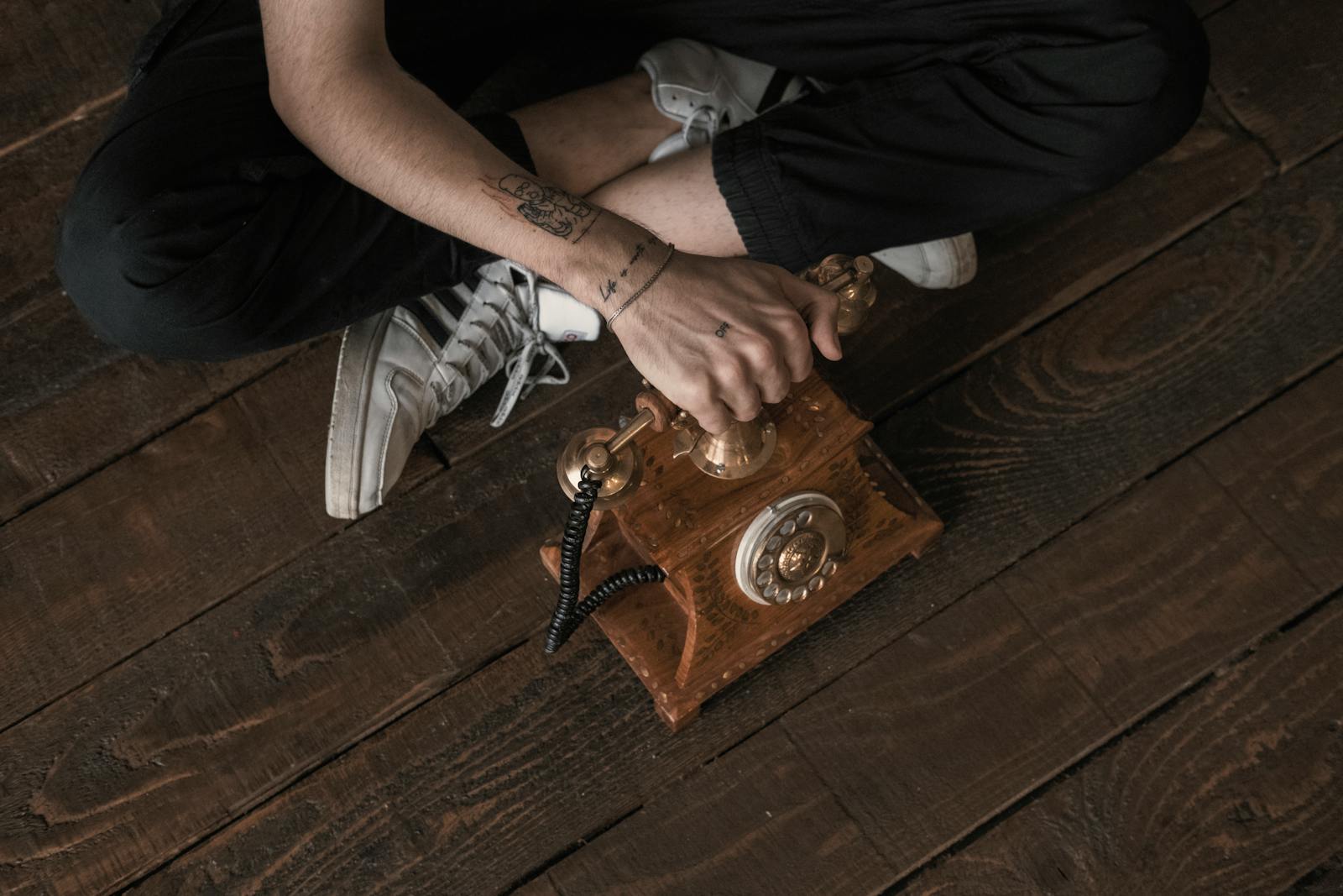 A tattooed hand dialing a vintage rotary phone on a wooden floor.