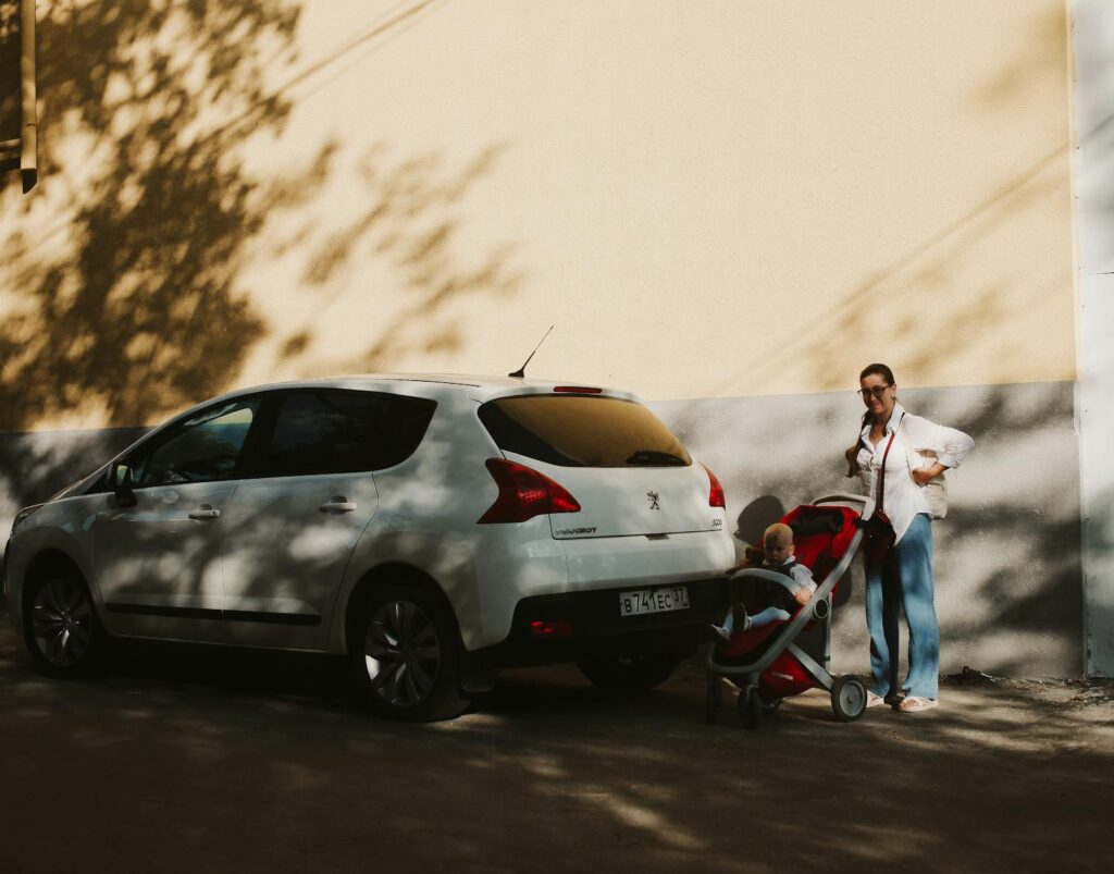 Mother with stroller and child beside a white car on a sunlit street.
