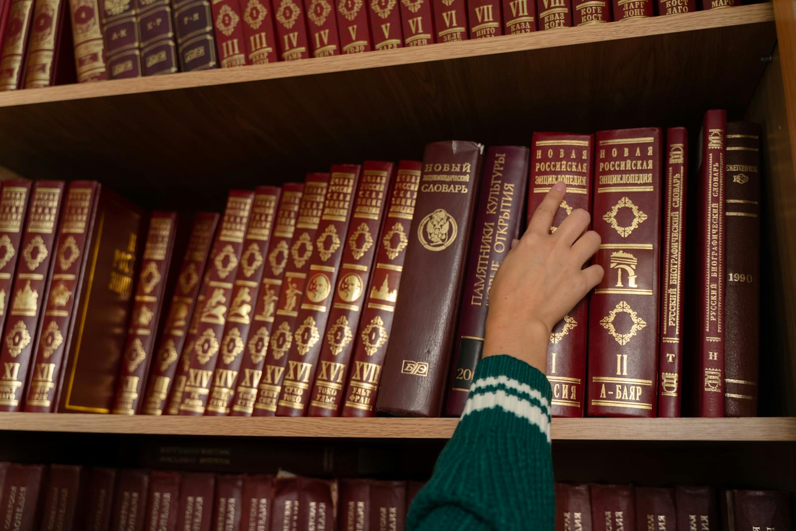 A person reaching for a red encyclopedia on a neatly organized bookshelf in a library.