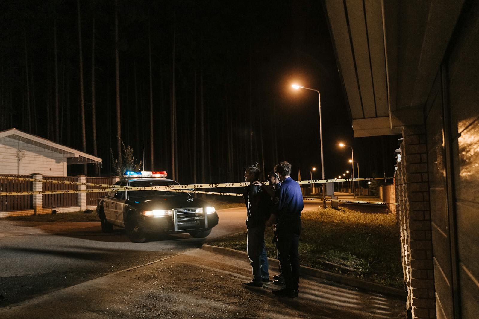 Police officers at a nighttime crime scene with a police car and caution tape under streetlights.