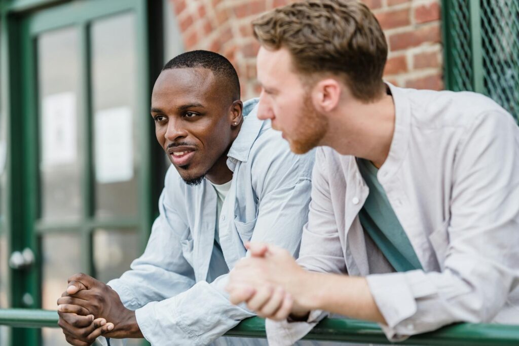 Two young men chatting and smiling while leaning on a railing outdoors.