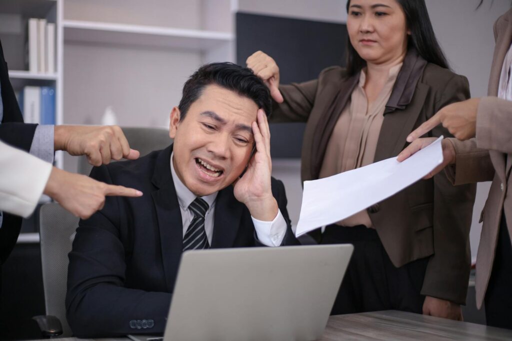 An Asian businessman looks stressed as colleagues point fingers and give him paperwork in a modern office.