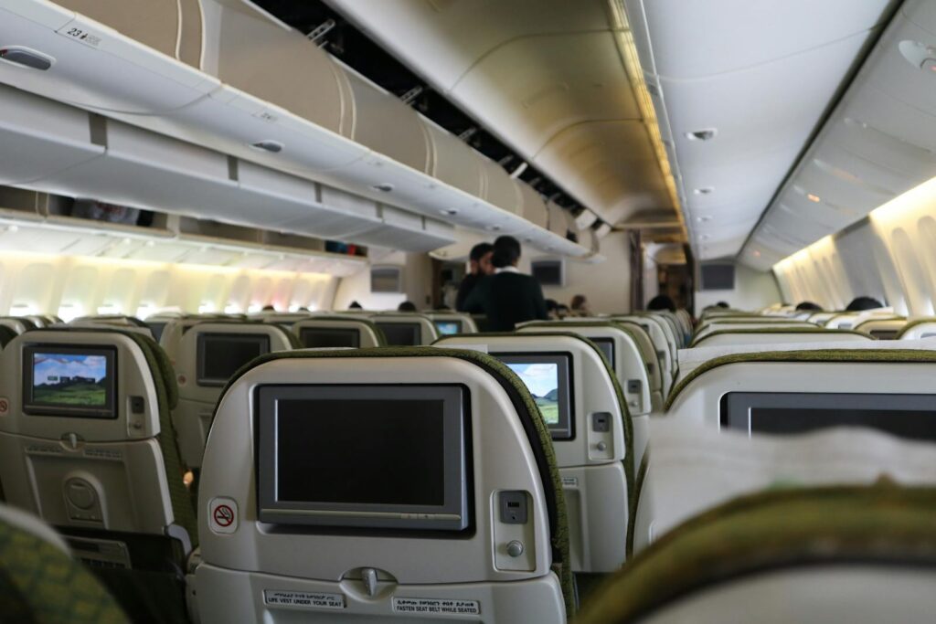 Interior view of an airplane cabin with seats and passengers, featuring screens and overhead compartments.