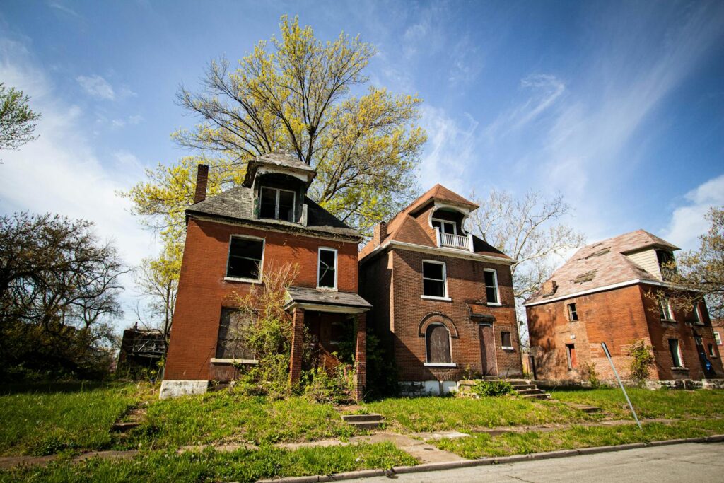 Three abandoned brick houses in a suburban neighborhood under a clear blue sky.