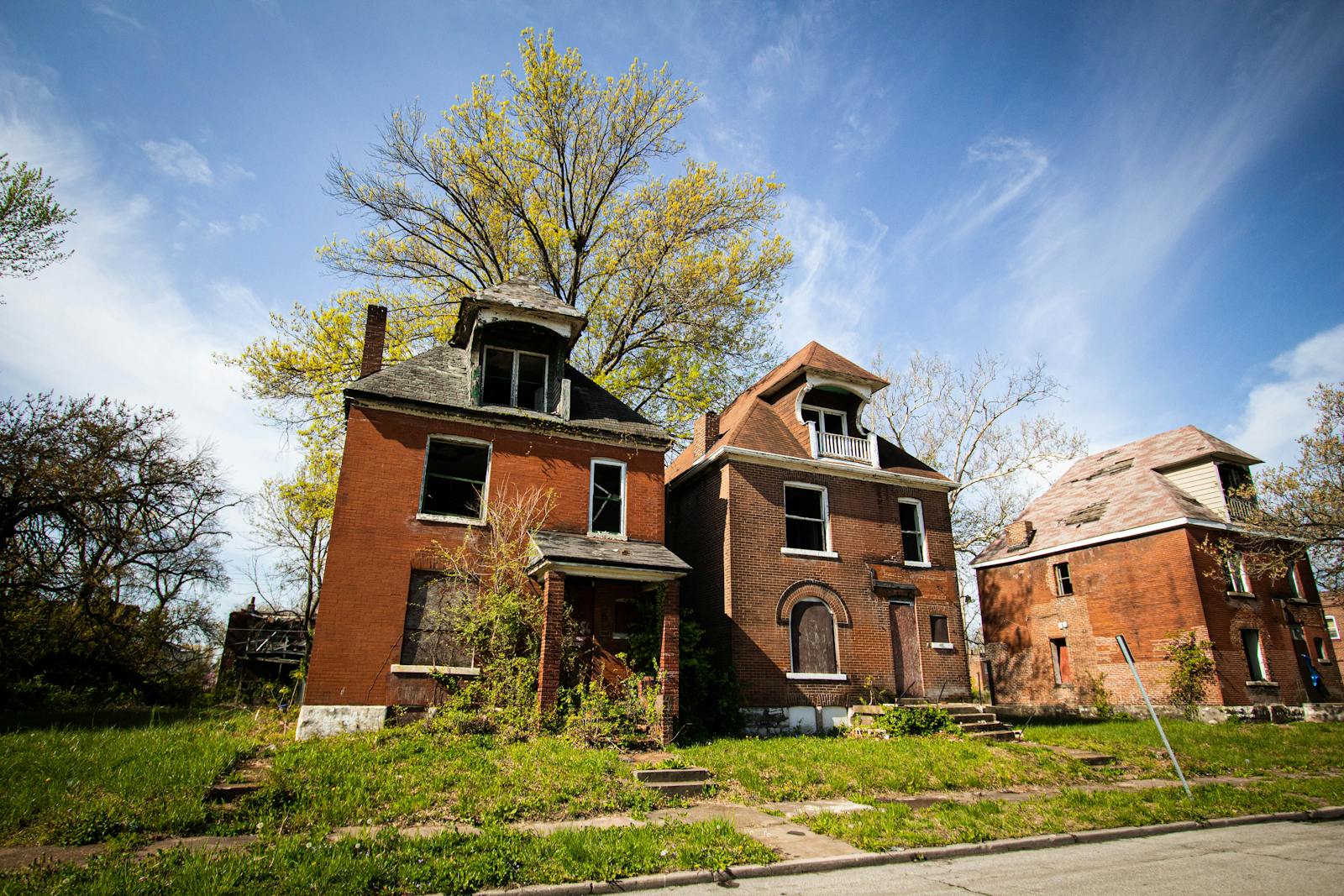Three abandoned brick houses in a suburban neighborhood under a clear blue sky.