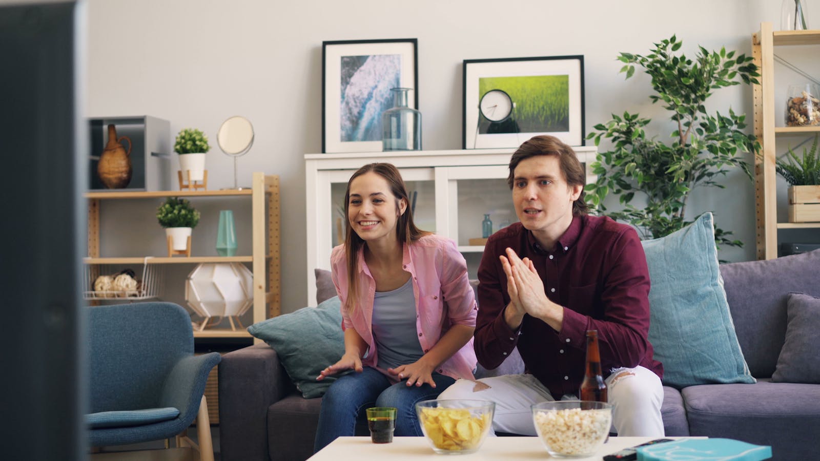 A young couple excitedly watching TV at home, enjoying snacks together in a lively living room setting.