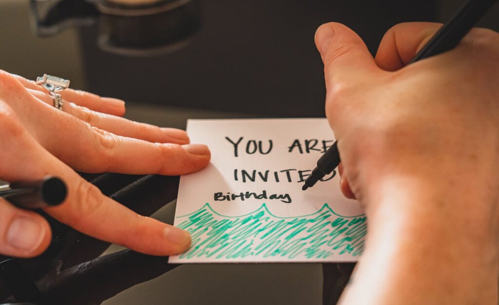 Hands writing a personalized birthday invitation card using a marker on paper.