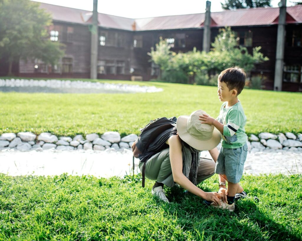 A mother assists her young son with his shoes on a sunny day in a grassy area.