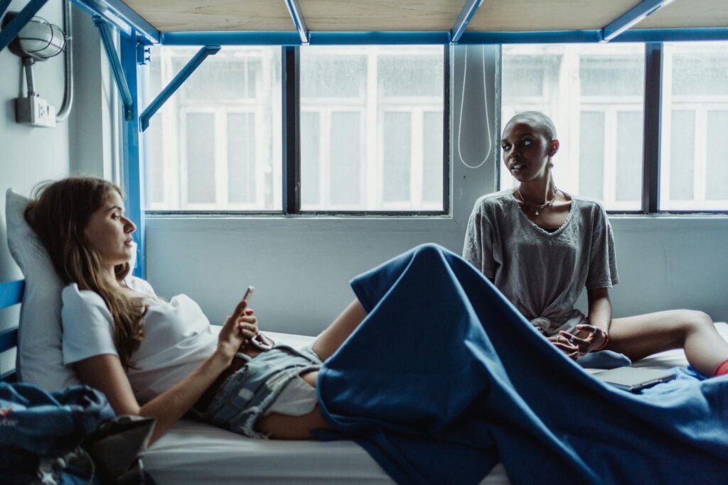 Two women sitting on a bunk bed, talking and relaxing in a hostel room.