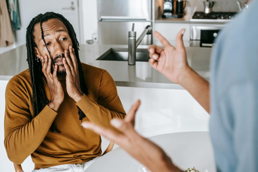 Young African American male sitting at table with hands on face and having conflict with female on kitchen