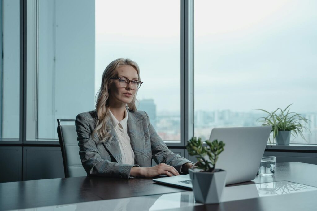 A confident businesswoman in a grey suit working on a laptop in a modern office setting.
