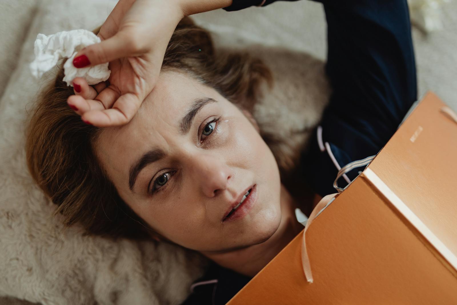 A woman lying on a bed, holding a tissue, showing an emotional expression.