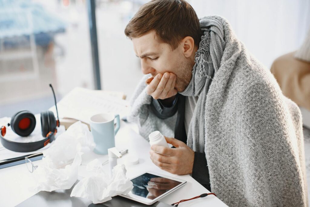 Man wrapped in blanket feeling unwell at home, surrounded by tissues and medication.