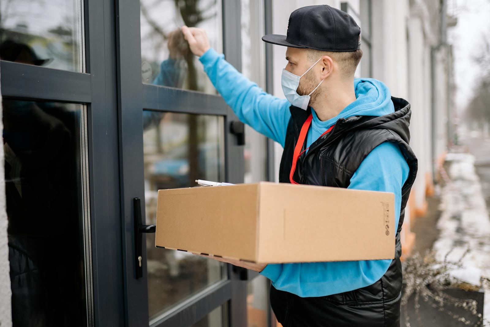 Male courier wearing a face mask delivers a package to a modern door, promoting contactless delivery.