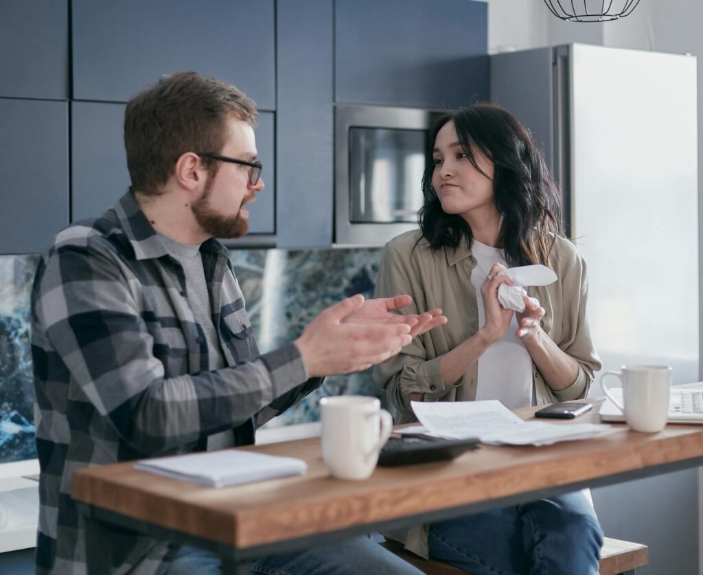 A couple sits at a kitchen table discussing bills, showcasing frustration and financial stress.