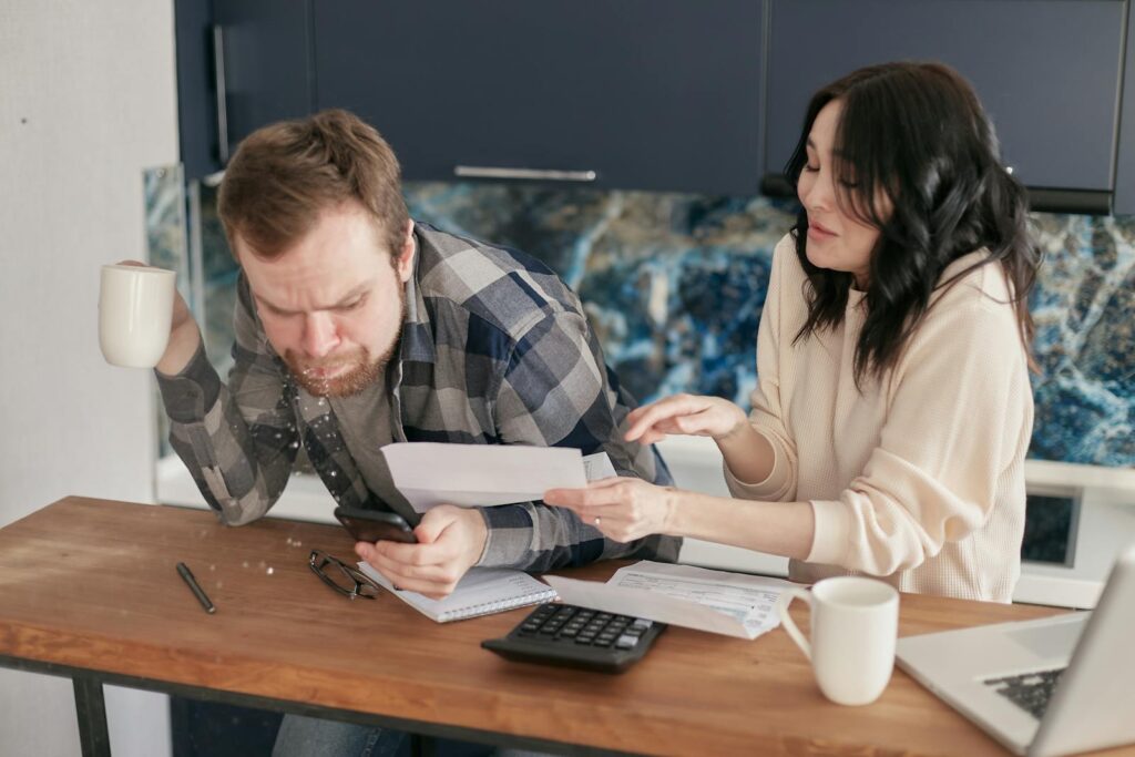 A couple sitting at a desk, reviewing bills and expenses with a calculator and laptop, looking worried.