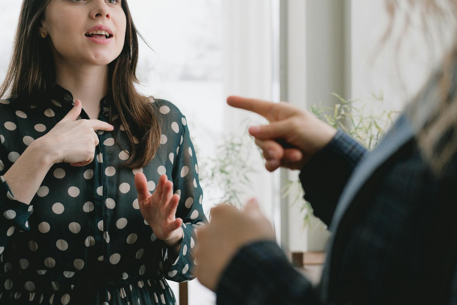 A young woman in a polka dot dress gestures expressively during a conversation indoors.