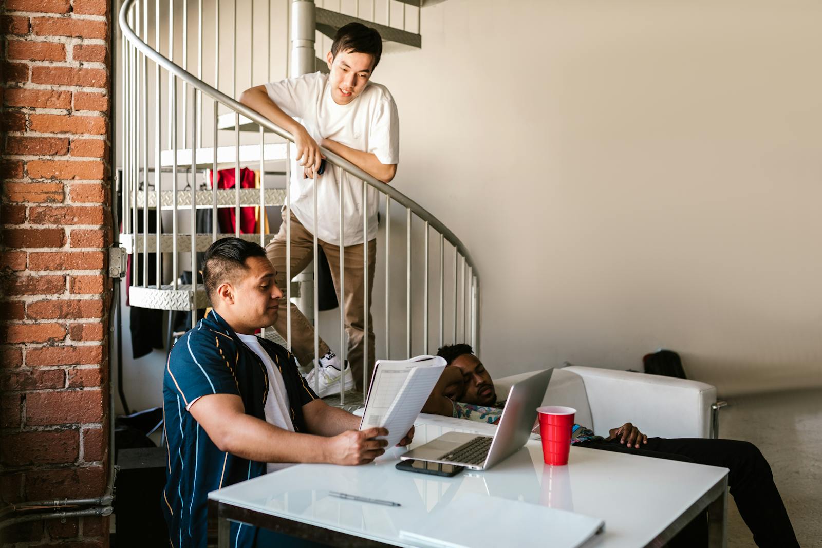 Young adults in an urban apartment discussing study materials around a spiral staircase.