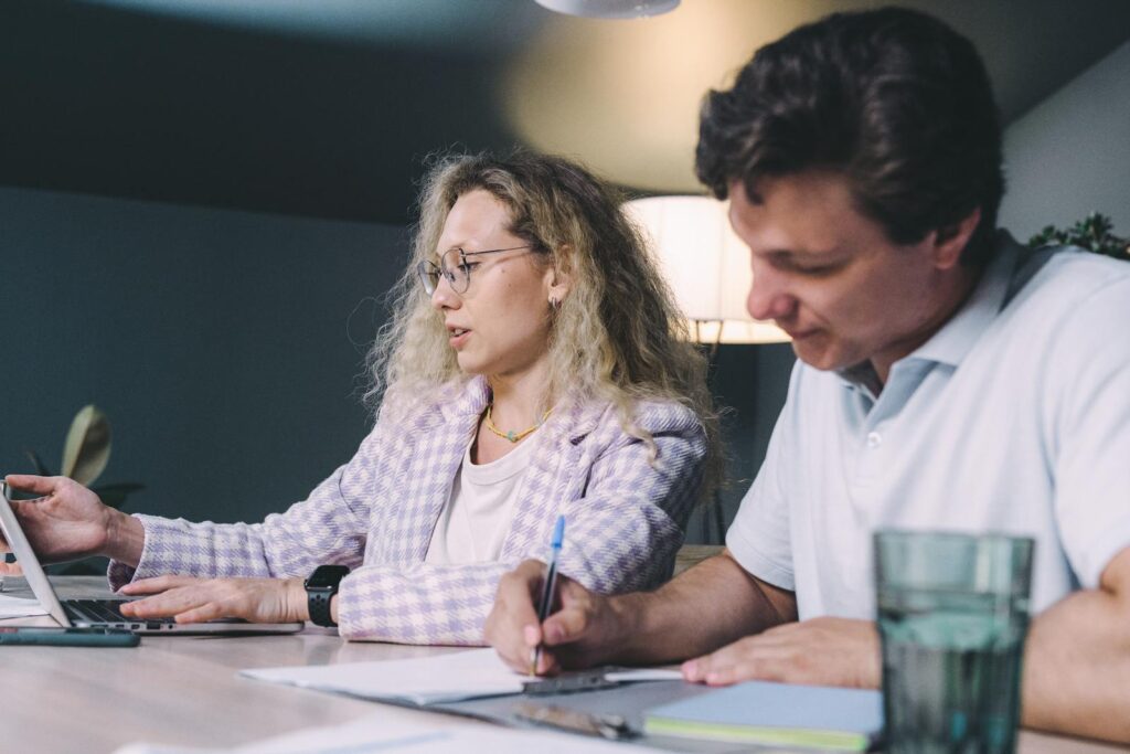 Two colleagues collaborating at a desk with laptops and papers in a modern office setting.