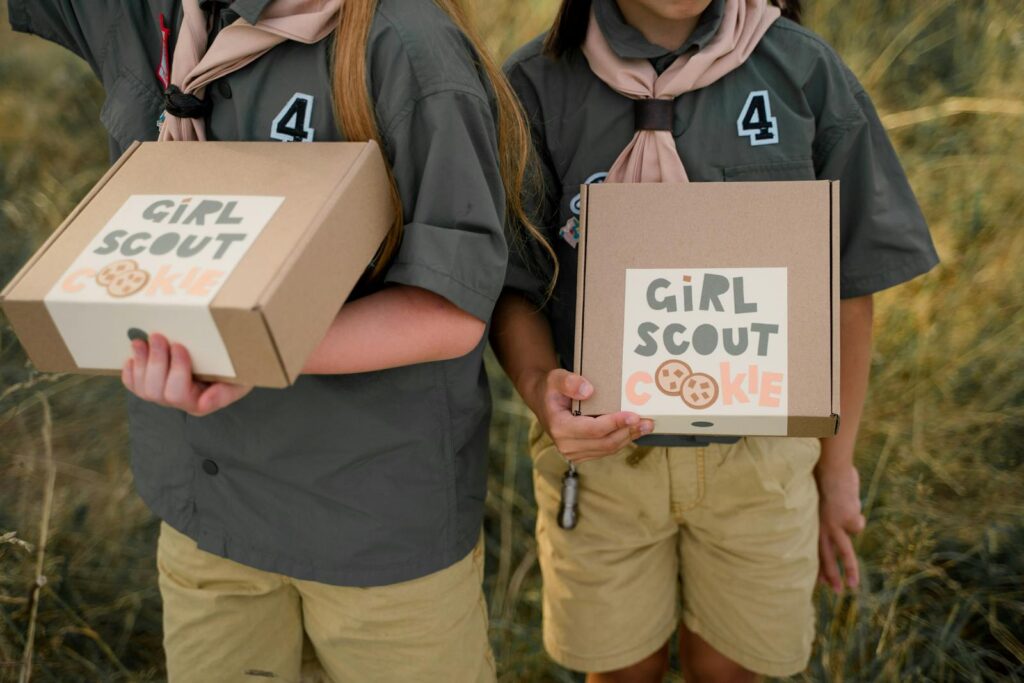 Two girl scouts holding cookie boxes with visible logos in an outdoor setting.