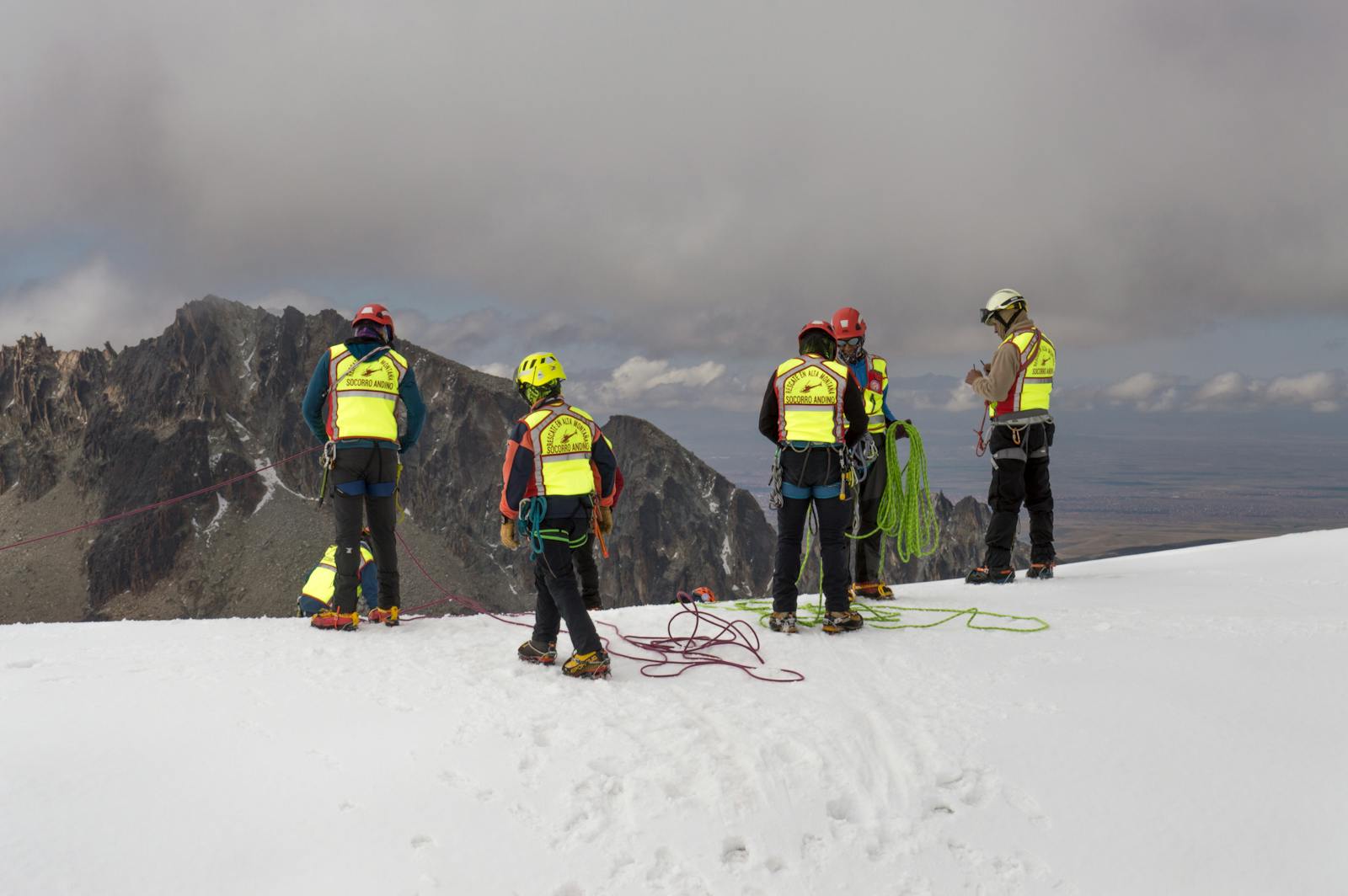 Group of mountain rescuers in safety vests and helmets preparing ropes in snowy terrain.
