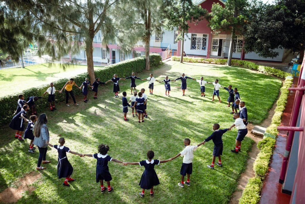 Group of children holding hands in a circle on a sunny schoolyard lawn.