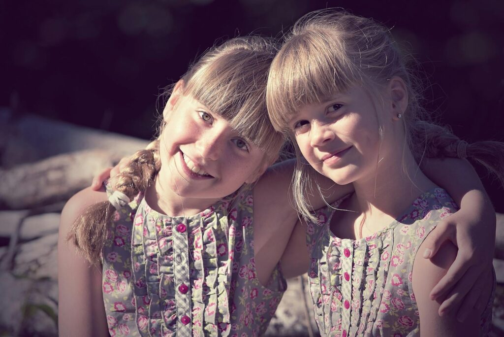 Two young sisters smile and embrace in matching floral dresses during a sunny day outdoors.