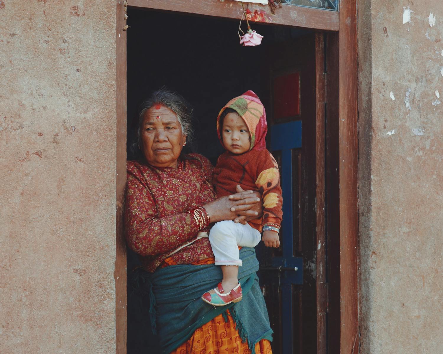 An elderly woman holding a child stands in a rustic doorway, capturing family and cultural essence.